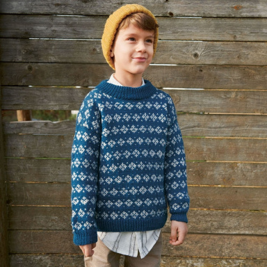 Sandnes Garn Harald Pullover für Kinder, getragen von einem Jungen mit gelbem Hut vor Holzlatten.