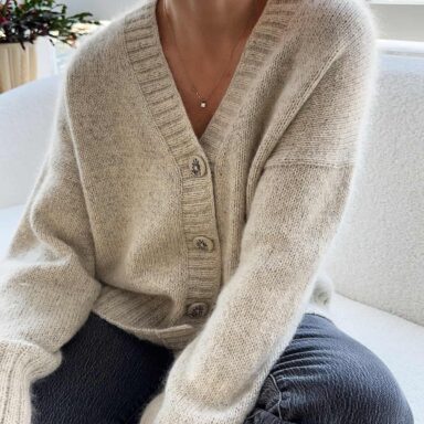 Weißer Cardigan aus Bio-Wolle, getragen von einer Frau, sitzt bequem auf einem Sofa vor einem Fenster mit natürlichem Li.