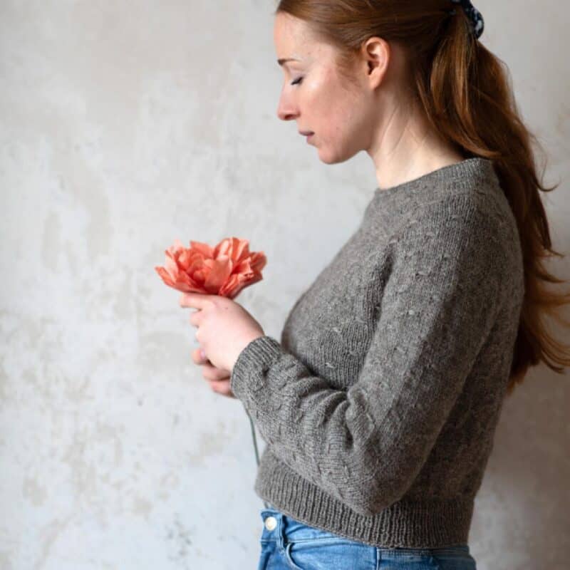 Frau hält eine orange Blume und trägt einen grauen Strickpullover, im Hintergrund eine helle Wand.